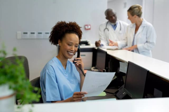 Photo of a female receptionist talking on the phone with a document in her hand, with two doctors having a conversation behind her