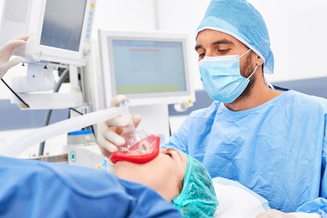 Photo of a male anesthesiologist administering anesthesia with a mask on the face of a patient, behind them are two medical equipment screens